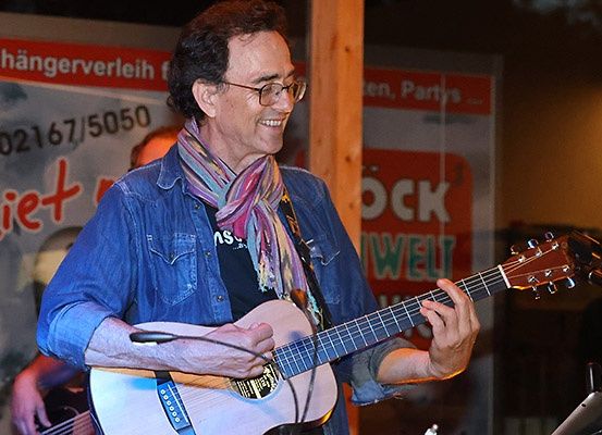 Ein Mann spielt Gitarre und lächelt. Er trägt eine Brille, einen Schal und eine Jeansjacke. Dahinter steht eine weitere Person, die ebenfalls Gitarre spielt. Im Hintergrund befindet sich ein Plakat mit Text.