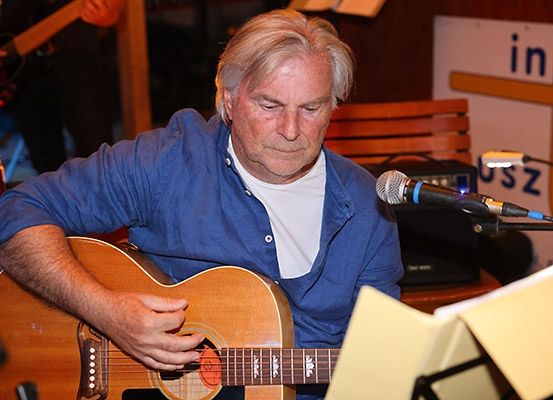 Ein Mann spielt eine Gitarre mit einem Mikrofon vor sich. Er schaut nach unten und zupft die Gitarre. Hinter ihm steht eine Holzbank.