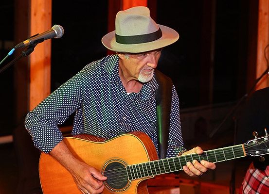 Ein älterer Mann mit Bart und Hut spielt Gitarre vor einem Mikrofon. Eine Frau in einem karierten Rock steht hinter ihm.