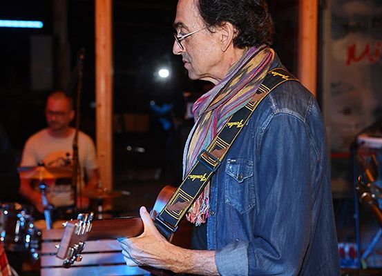 Ein älterer Mann spielt Gitarre und schaut nach unten. Hinter ihm spielt ein Mann die Drums. Der Hintergrund ist verschwommen mit einem Licht und einem Schild mit der Aufschrift 'Mic'.
