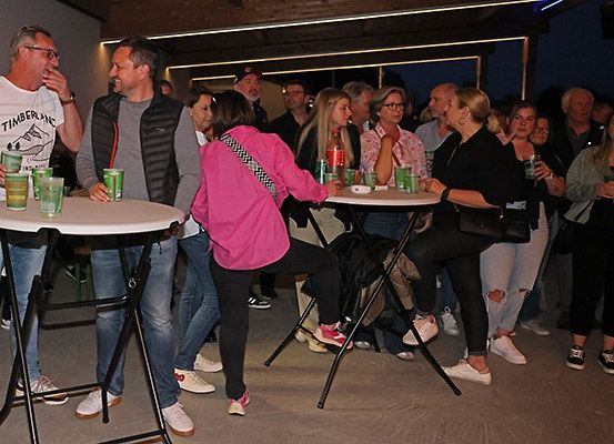 Eine Gruppe von Menschen steht und sitzt um Tische herum, wahrscheinlich auf einer Party, mit Getränken auf den Tischen und einigen, die in der Nähe der Wand stehen.