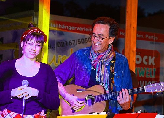 Eine Frau in einem lila Shirt hält ein Mikrofon und ein Mann in einer Jeansjacke spielt Gitarre. Sie treten auf einer Bühne auf.