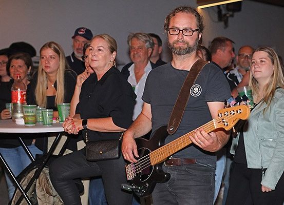 Ein Mann spielt eine Bassgitarre in einem überfüllten Raum, umgeben von einem Publikum. Er steht vor einem Tisch mit Getränken. Eine Frau neben ihm beobachtet ihn beim Spielen.