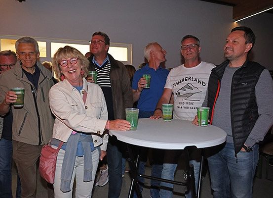 Eine Gruppe Erwachsener steht um einen Tisch herum und hält grüne Becher in der Hand. Eine Frau lächelt und hält einen Becher in der Hand. Hinter ihr trägt ein Mann eine Jacke mit einem weißen T-Shirt, auf dem Timberland steht. Ein weiterer Mann in einer Weste steht rechts.