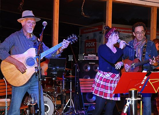 Drei Musiker treten nachts auf einer Bühne auf, einer spielt Gitarre und singt, ein anderer singt und spielt Gitarre, und ein Schlagzeug ist im Hintergrund zu sehen.