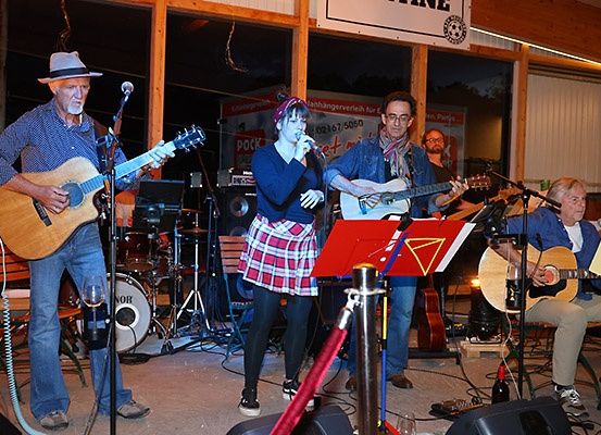Eine Musikerband tritt auf einer Bühne auf. Eine Frau singt, während ein Mann Gitarre spielt. Zwei andere Männer spielen Gitarre und Schlagzeug. Die Umgebung umfasst Lautsprecher und Musikinstrumente.