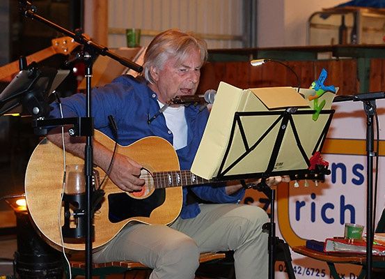 Ein Mann sitzt auf einem Stuhl und spielt Gitarre. Er schaut auf das Notenblatt, das auf einem Notenständer vor ihm steht. Er hält die Gitarre mit der linken Hand.
