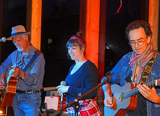 Drei Musiker treten auf einer Bühne auf. Ein Mann links spielt Gitarre, ein anderer rechts ebenfalls Gitarre, und eine Frau in der Mitte singt in ein Mikrofon. Sie stehen unter blauen und orangefarbenen Bühnenlichtern.