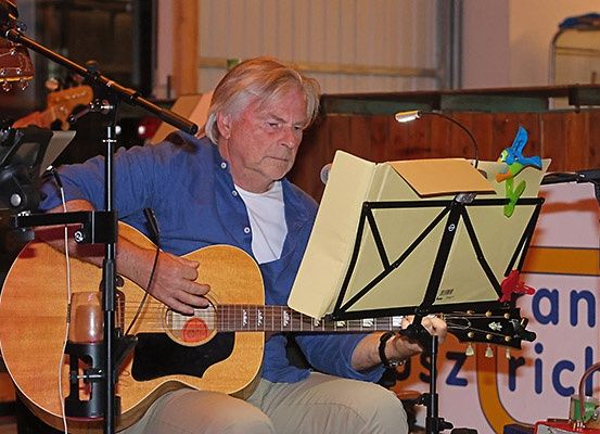 Ein älterer Mann sitzt und spielt Gitarre. Vor ihm steht ein Notenständer mit Notenblättern. Er hat ein Mikrofon auf einem Ständer und einen Glas auf der Gitarre. Er schaut auf die Notenblätter. Hinter ihm ist eine Holzwand mit einem Plakat.