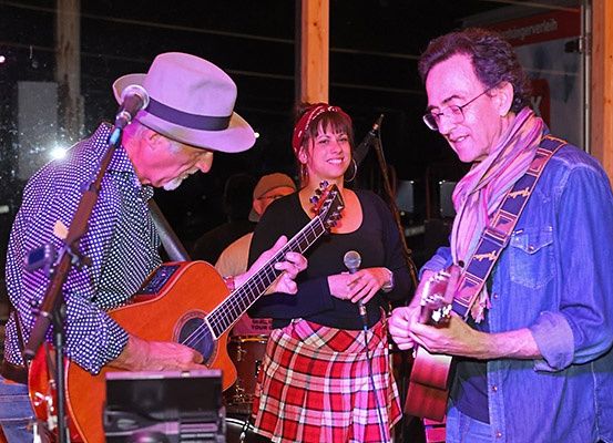 Drei Menschen spielen Musik. Ein Mann auf der linken Seite spielt Gitarre und singt. Die Frau in der Mitte spielt ebenfalls Gitarre und singt. Ein anderer Mann auf der rechten Seite spielt Gitarre. Alle lächeln.