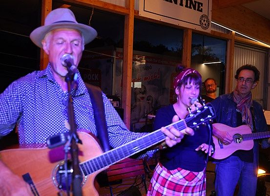 Eine Band tritt mit Mikrofon und Gitarren auf der Bühne auf, unter einem Schild mit der Aufschrift 'entine'. Eine Frau in einem Karorock singt, ein Mann spielt Gitarre und ein anderer spielt Gitarre hinter ihr. Ein weiterer Mann steht hinter ihnen.