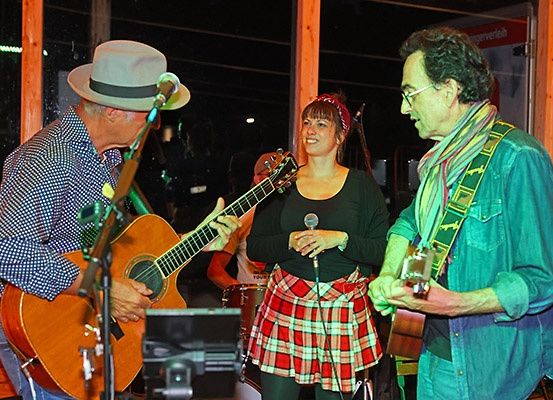 Drei Musiker treten bei Nacht auf der Bühne auf. Ein Mann in einem Hut spielt Gitarre, ein anderer mit einem Schal spielt Gitarre und eine Frau in einem karierten Rock hält ein Mikrofon. Dahinter spielt ein Schlagzeuger.