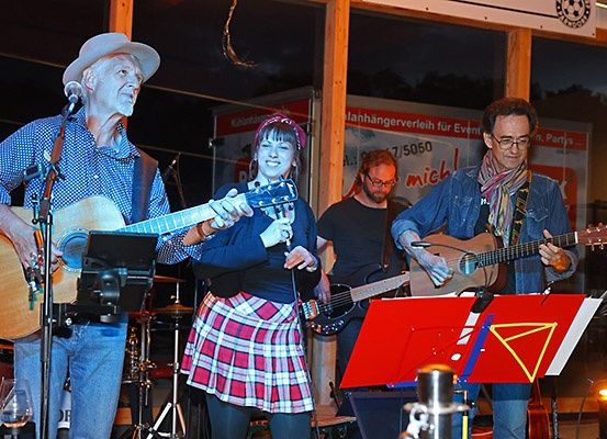 Eine Band mit vier Musikern tritt nachts auf einer Bühne auf. Ein Mann spielt Gitarre, während eine Frau singt. Ein weiterer Mann spielt Gitarre und ein Mann spielt Schlagzeug. Vor ihnen steht ein Notenständer. Hinter ihnen befindet sich eine Glaswand mit einem Schild.