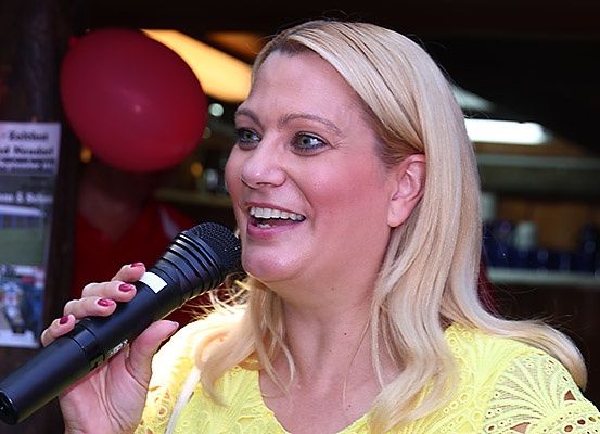 Eine Frau mit blondem Haar hält ein Mikrofon, lächelt und spricht. Sie trägt ein gelbes Oberteil. Hinter ihr befinden sich zwei rote Luftballons und ein unscharfer Hintergrund.