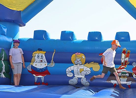 Zwei Kinder spielen in einem blauen aufblasbaren Schloss. Eines läuft und das andere steht. Das aufblasbare Schloss hat Comicfiguren darauf.