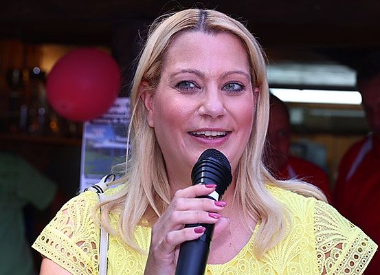 Eine Frau mit blonden Haaren trägt ein gelbes Oberteil und hält ein Mikrofon in der Hand. Sie scheint sich in einem Restaurant zu befinden, wobei ein roter Ballon im Hintergrund zu sehen ist.