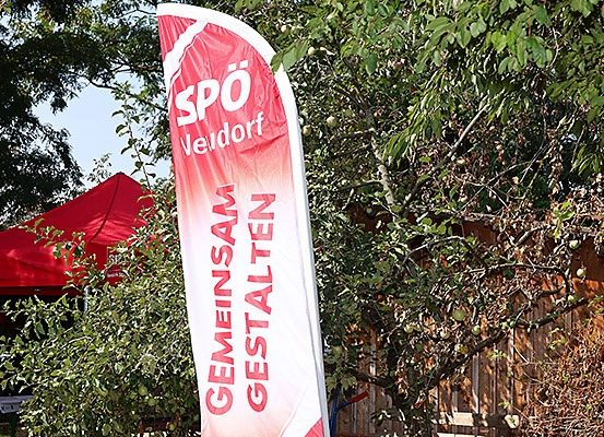 Ein roter Banner mit weißem Text steht vor einem roten Zelt und einem Holzzaun mit Pflanzen und Bäumen.