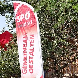 Ein roter Banner mit weißem Text steht vor einem roten Zelt und einem Holzzaun mit Pflanzen und Bäumen.