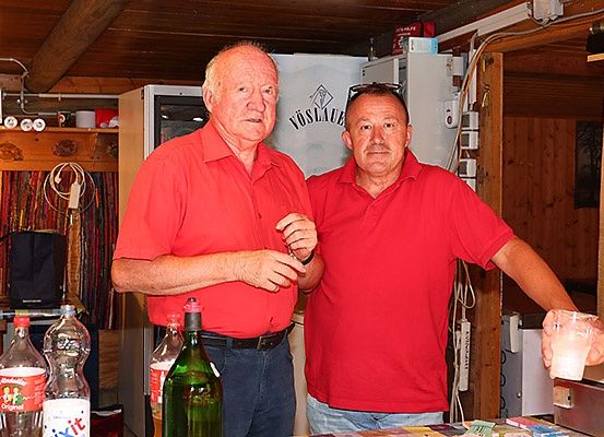 Zwei Männer in roten Hemden stehen in einem Raum mit einem Tisch und Flaschen. Einer hält ein Getränk und der andere hat eine Brille auf dem Kopf. Ein Kühlschrank und Regale mit Gegenständen sind im Hintergrund.