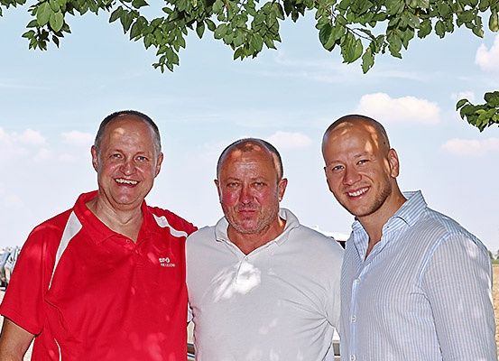 Drei Männer stehen zusammen unter einem Baum und lächeln für ein Foto. Der Mann links trägt ein rotes Shirt, der in der Mitte ein weißes Polohemd und der rechts ein hellblau gestreiftes Hemd. Sie stehen auf einem Feld mit geparkten Autos in der Ferne.