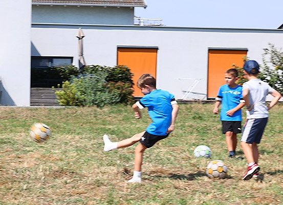 Drei Jungen spielen Fußball auf einem Rasenfeld, einer tritt den Ball, während die anderen beiden zuschauen. Ein Haus mit orangefarbenen Türen ist im Hintergrund.