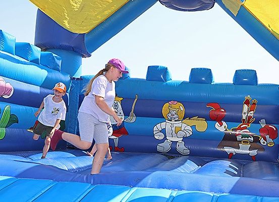 Zwei Kinder spielen in einem blauen aufblasbaren Hüpfburg mit Zeichentrickfiguren darauf. Ein Kind springt, während das andere zuschaut.