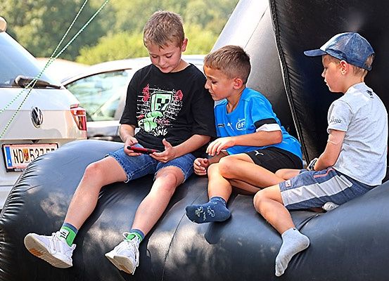 Drei Jungen sitzen auf einem großen aufblasbaren schwarzen Ball. Einer hält ein Handy, während die anderen es betrachten. Sie sind draußen in der Nähe geparkter Autos und Bäume.