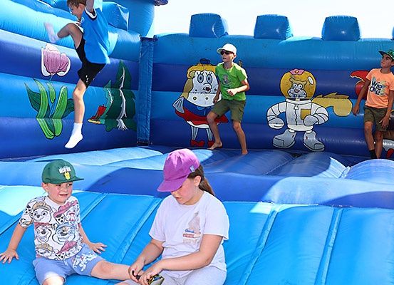 Mehrere Kinder spielen auf einem blauen aufblasbaren Sprunghaus, das mit Zeichentrickfiguren verziert ist. Ein Junge springt, während andere sitzen und herumstehen.