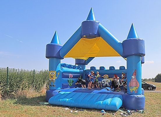 Mehrere Kinder springen in einem großen blauen aufblasbaren Schloss mit gelber Einfassung, umgeben von Gras und einem Zaun. Ein Auto ist in der Nähe geparkt.