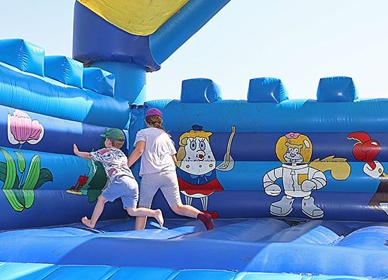 Zwei Kinder spielen in einem blauen aufblasbaren Sprunghaus mit gezeichneten Zeichentrickfiguren darauf, unter einem klaren Himmel.