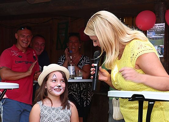 Eine Frau spricht in ein Mikrofon, während ein junges Mädchen mit Katzengesichtsbemalung zuhört. Dahinter lachen zwei Männer und eine Frau, und ein Banner hängt im Hintergrund.