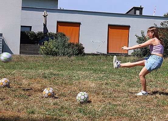 Ein Mädchen tritt in einem Grasfeld mit einem Haus im Hintergrund Fußballbälle.