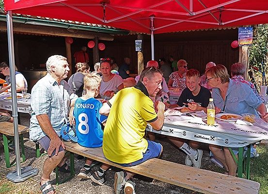 Mehrere Personen sitzen unter einem roten Baldachin an Tischen und essen. Ein Mann trägt ein gelbes Shirt mit der Nummer 8. Ein Junge in einem blauen Shirt sitzt neben ihm. Eine Frau lächelt in der Nähe.