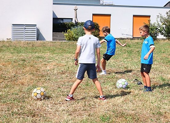 Drei Jungen spielen Fußball auf einem Rasenfeld vor einem Haus mit orangefarbenen Fensterläden. Ein Junge läuft, während die anderen ihn beobachten.
