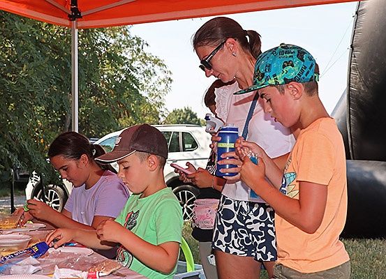 Eine Gruppe von Kindern und Erwachsenen versammelt sich unter einem Zelt und bereitet Essen zu. Die Erwachsenen beaufsichtigen die Kinder, die Besteck und Essen verwenden.