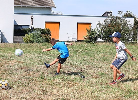 Zwei Jungen spielen Fußball auf einem Grasfeld vor einem modernen weißen Haus mit orangefarbenen Rollläden.