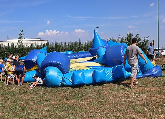 Kinder spielen auf einem blauen aufblasbaren Hüpfburg im Feld, ein Mann steht in der Nähe und ein anderer Mann hält einen Eimer. Dahinter sind Büsche, ein weißes Auto und ein Gebäude.