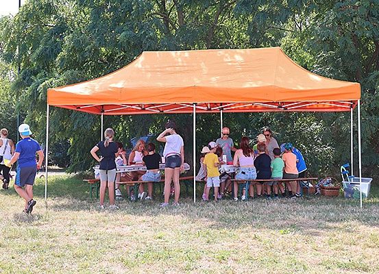 Eine Gruppe von Menschen sitzt unter einem orangefarbenen Baldachin in einem Park an Picknicktischen. Einige tragen Helme.