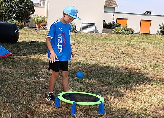 Ein Junge in einem blauen Shirt und einer Kappe spielt mit einem blauen Ball im Hinterhof, in der Nähe eines Trampolins und einer aufblasbaren Hürde.
