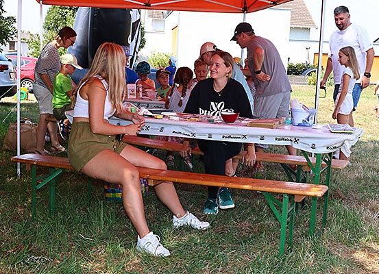 Eine Gruppe von Menschen, einschließlich Erwachsener und Kindern, versammelt sich um einen Picknicktisch unter einem Zelt. Sie essen und unterhalten sich. Eine Frau in einem weißen Tanktop und Shorts sitzt am Tisch.
