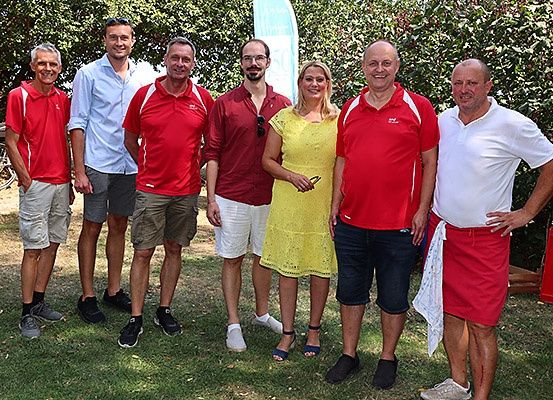 Eine Gruppe von Menschen steht auf einem Rasenfeld, lächelt und posiert für ein Foto. Einige tragen rote Poloshirts, andere weiße. Eine Frau in einem gelben Kleid steht in der Mitte. Hinter ihnen stehen Fahrräder und eine Fahne.