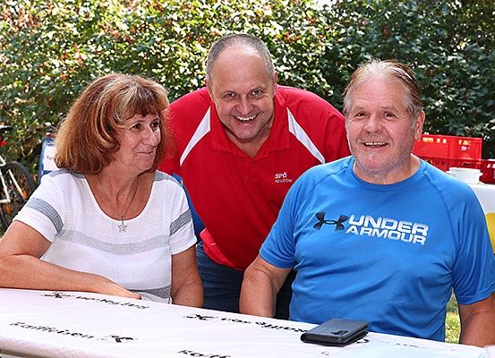 Drei Menschen lächeln für ein Foto an einem Tisch in einer Außenumgebung. Eine Frau links, ein Mann in einem roten Shirt in der Mitte und ein Mann in einem blauen Shirt rechts.