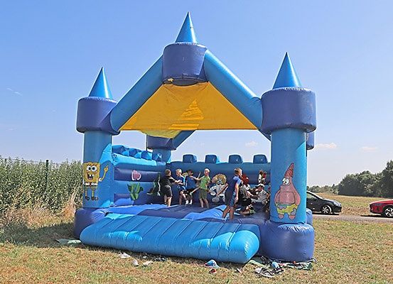 Eine Gruppe von Kindern spielt in einem blauen aufblasbaren Schloss mit gelbem Dach, das Cartoon-Charaktere zeigt. Sie sind von trockenem Gras umgeben, und zwei Autos sind im Hintergrund geparkt.