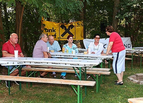 Fünf Personen sitzen an einem Tisch mit Getränken und Essen in einem Park, mit einer Frau in der Nähe und einem Banner im Hintergrund.