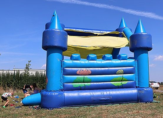 Ein blauer aufblasbarer Hüpfburg mit einem gelben Baldachin und Blumendekorationen steht auf einem Rasenfeld. Kinder spielen darum herum unter einem klaren blauen Himmel.