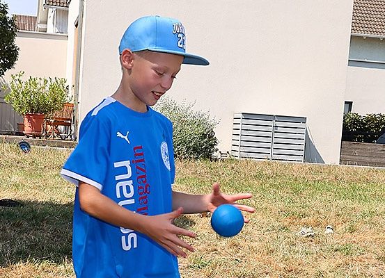 Ein Junge in einem blauen Hut und T-Shirt spielt mit einem blauen Ball im Garten.