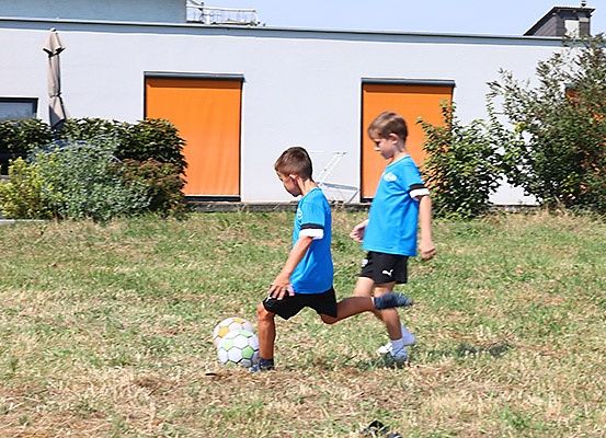 Zwei Jungen spielen Fußball auf einem Rasenfeld vor einem Gebäude mit orangefarbenen Türen.