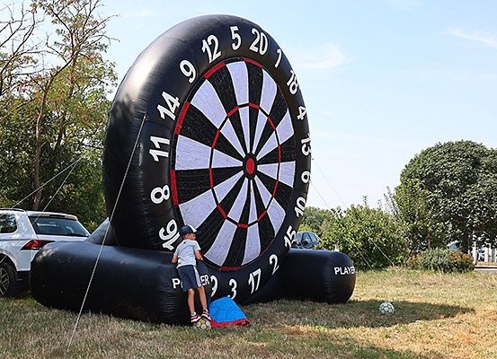 Ein junger Junge steht neben einem riesigen aufblasbaren Dartboard auf einem Rasenfeld, mit einem Auto und Bäumen im Hintergrund.