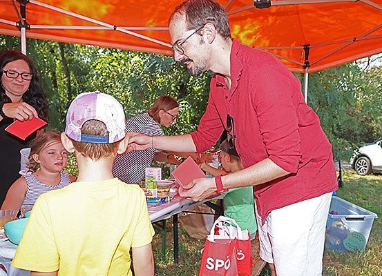 Ein Mann in Rot verteilt Gegenstände an ein Kind in einem gelben Shirt bei einer Veranstaltung im Freien.