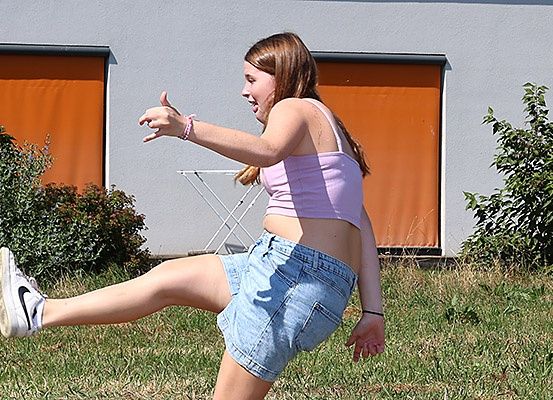 Eine Frau in Jeansshorts und einem rosa Tanktop tritt einen Ball auf einem Rasen mit einem orangefarbenen Gebäude im Hintergrund.